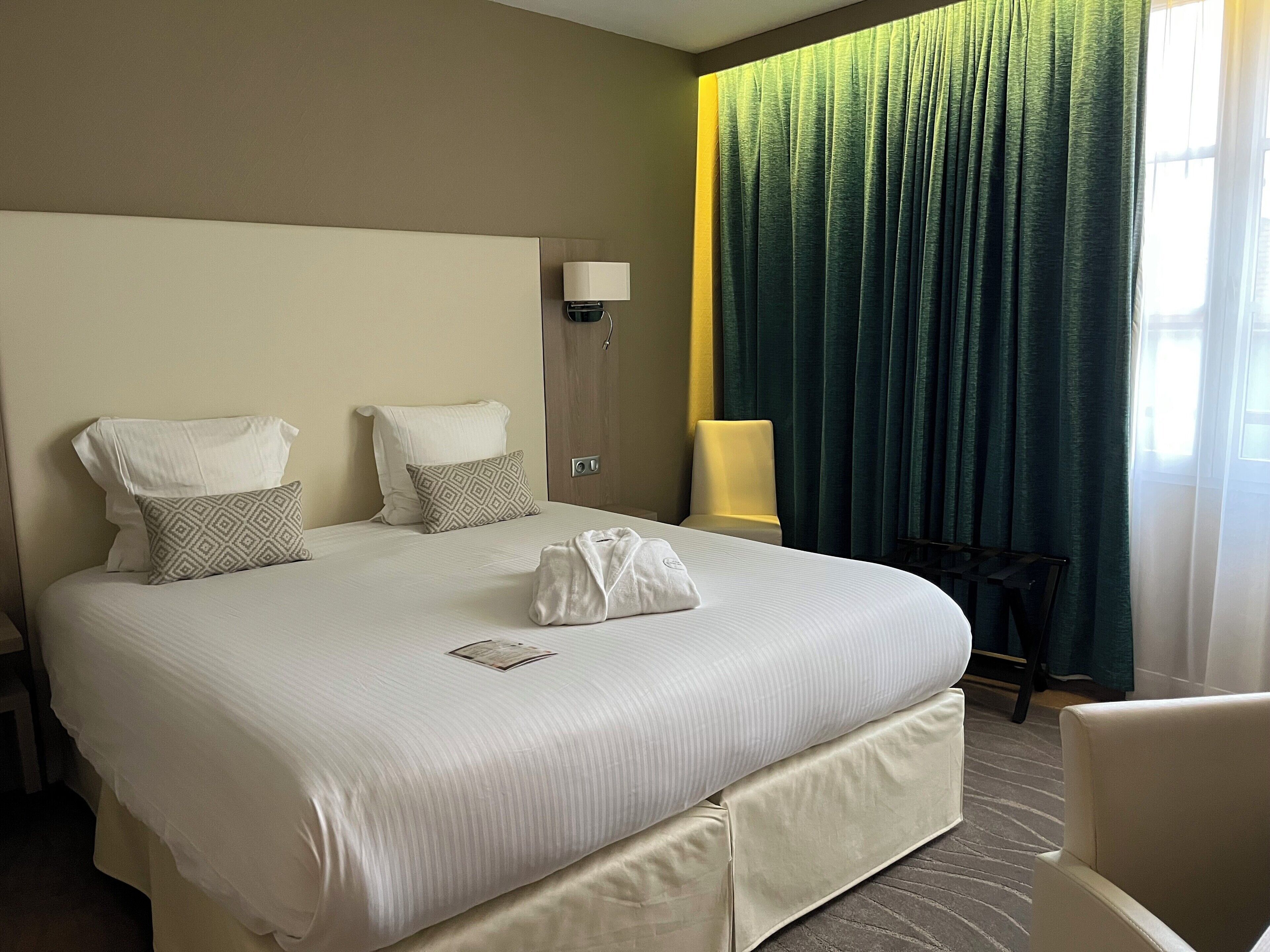 A hotel room with a large bed, two pillows, a bedside lamp, a chair, and a window with curtains.
