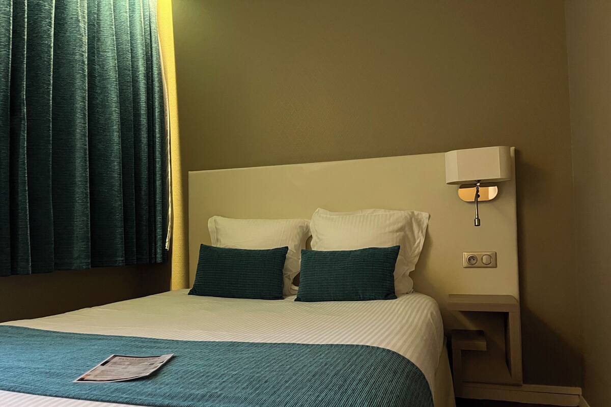 A hotel room with a bed, two pillows, a bedside table with a lamp, and a window with curtains.