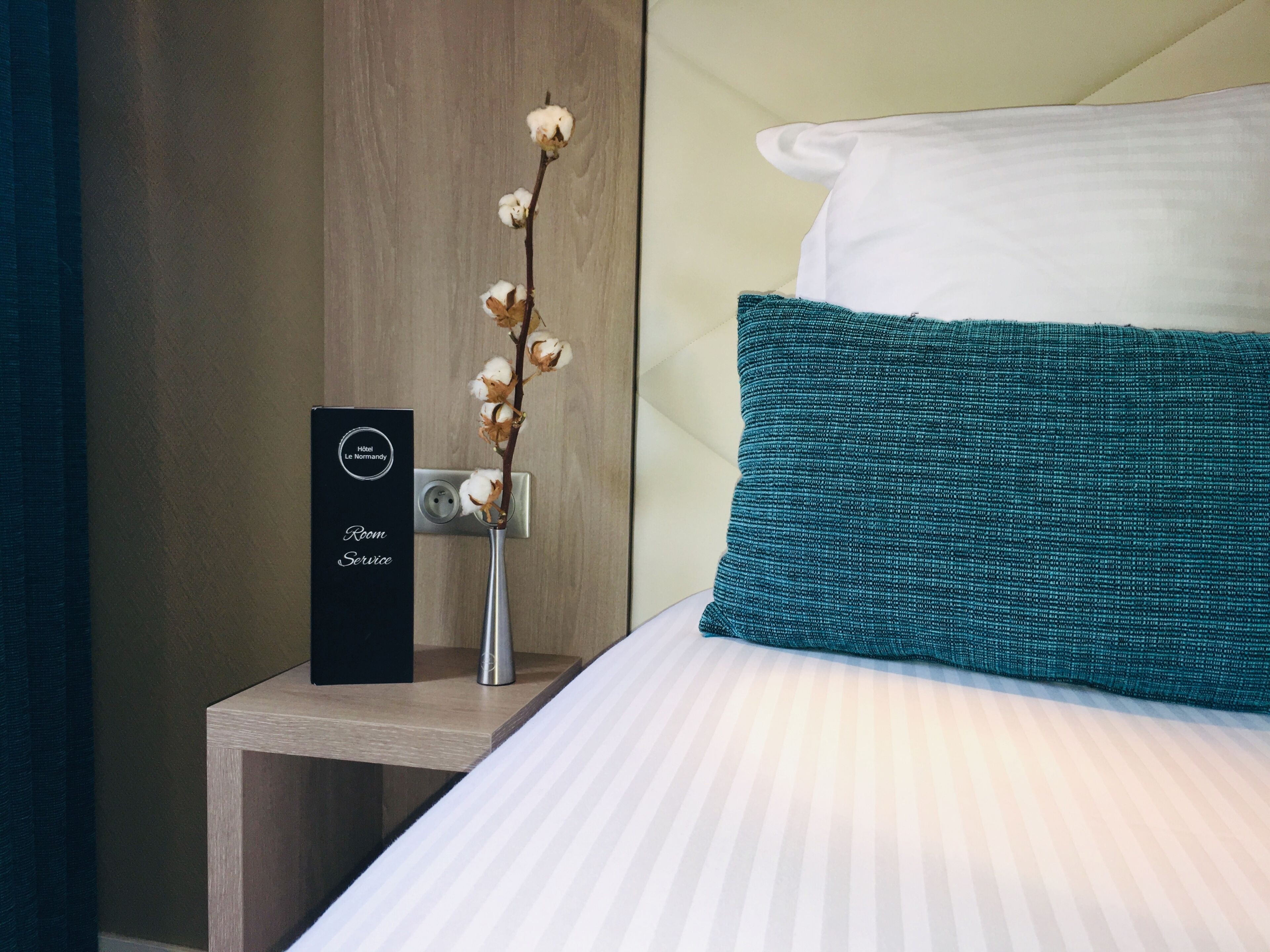 A neatly made bed with a teal pillow and a white pillow, a small bedside table with a vase of cotton flowers, and a black sign indicating room service.