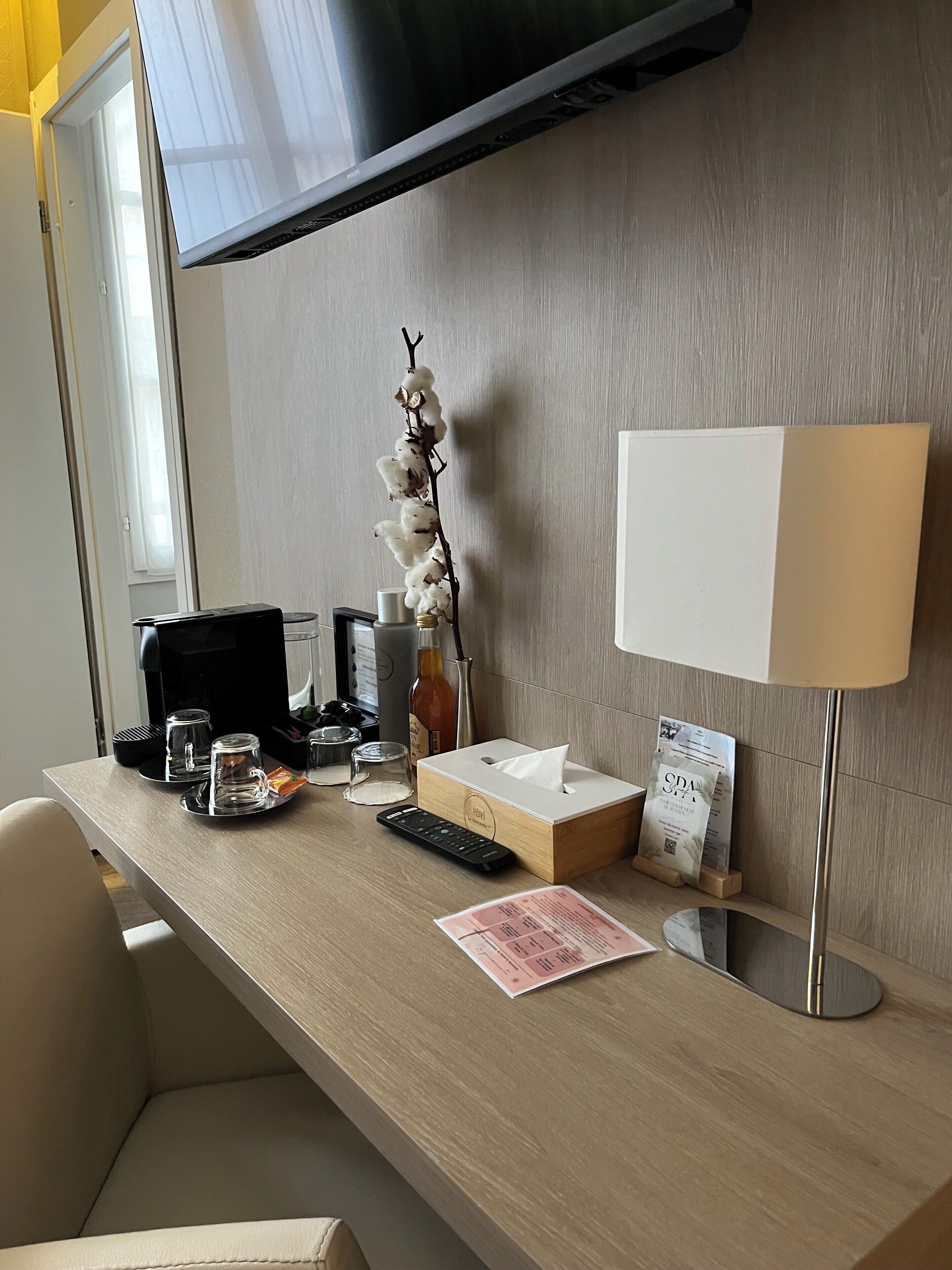 A hotel room with a wooden desk, a flat-screen TV mounted on the wall, a bedside table with a lamp, a tissue box, a remote control, and a menu on the table.