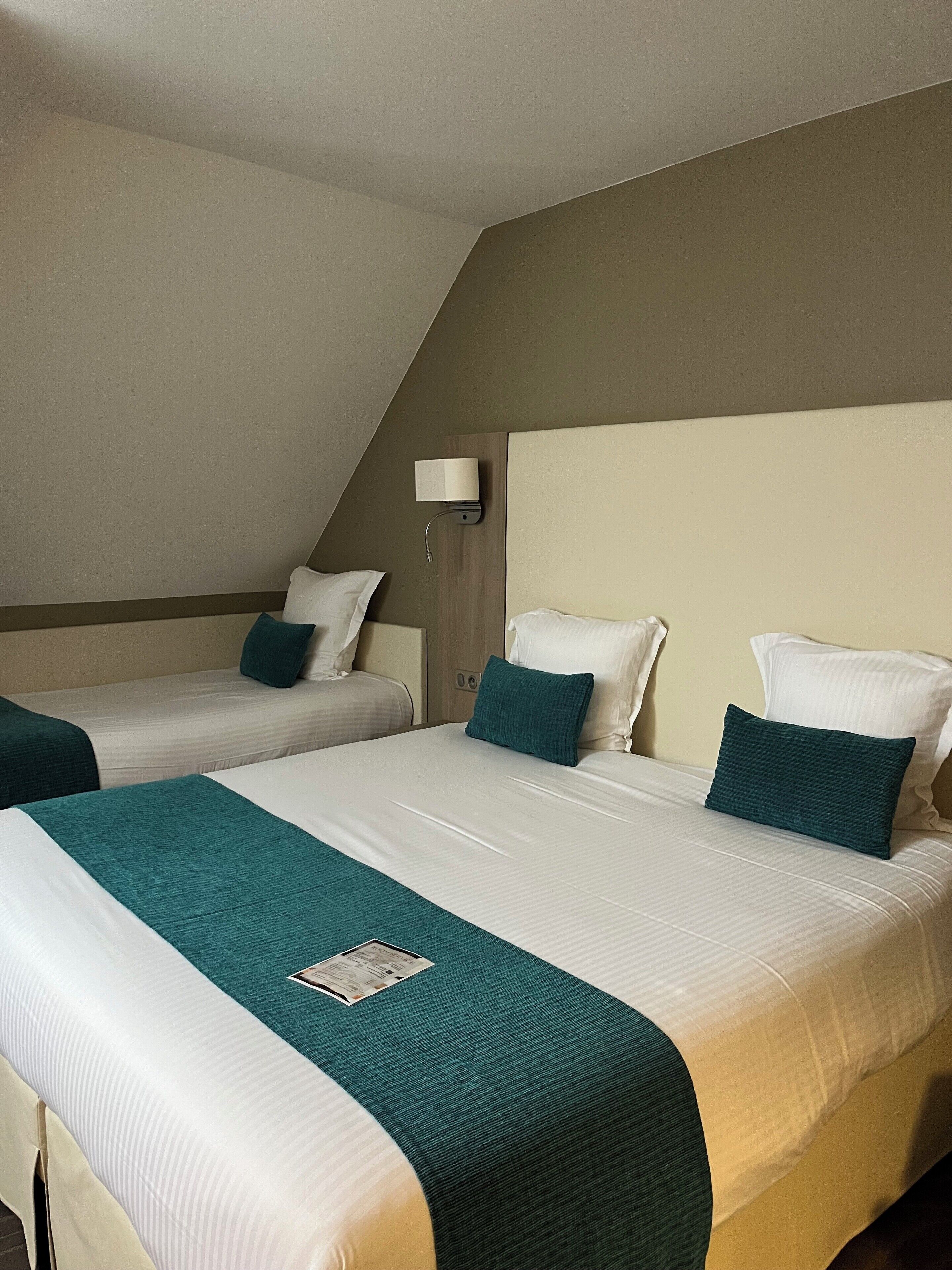 Quarto de hotel com duas camas de solteiro e uma cama maior. Cada cama possui um edredom branco e almofadas decorativas azul-turquesa. Há uma luminária fixada na parede acima da cama maior.