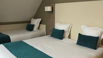 Quarto de hotel com duas camas de solteiro e uma cama maior. Cada cama possui um edredom branco e almofadas decorativas azul-turquesa. Há uma luminária fixada na parede acima da cama maior.
