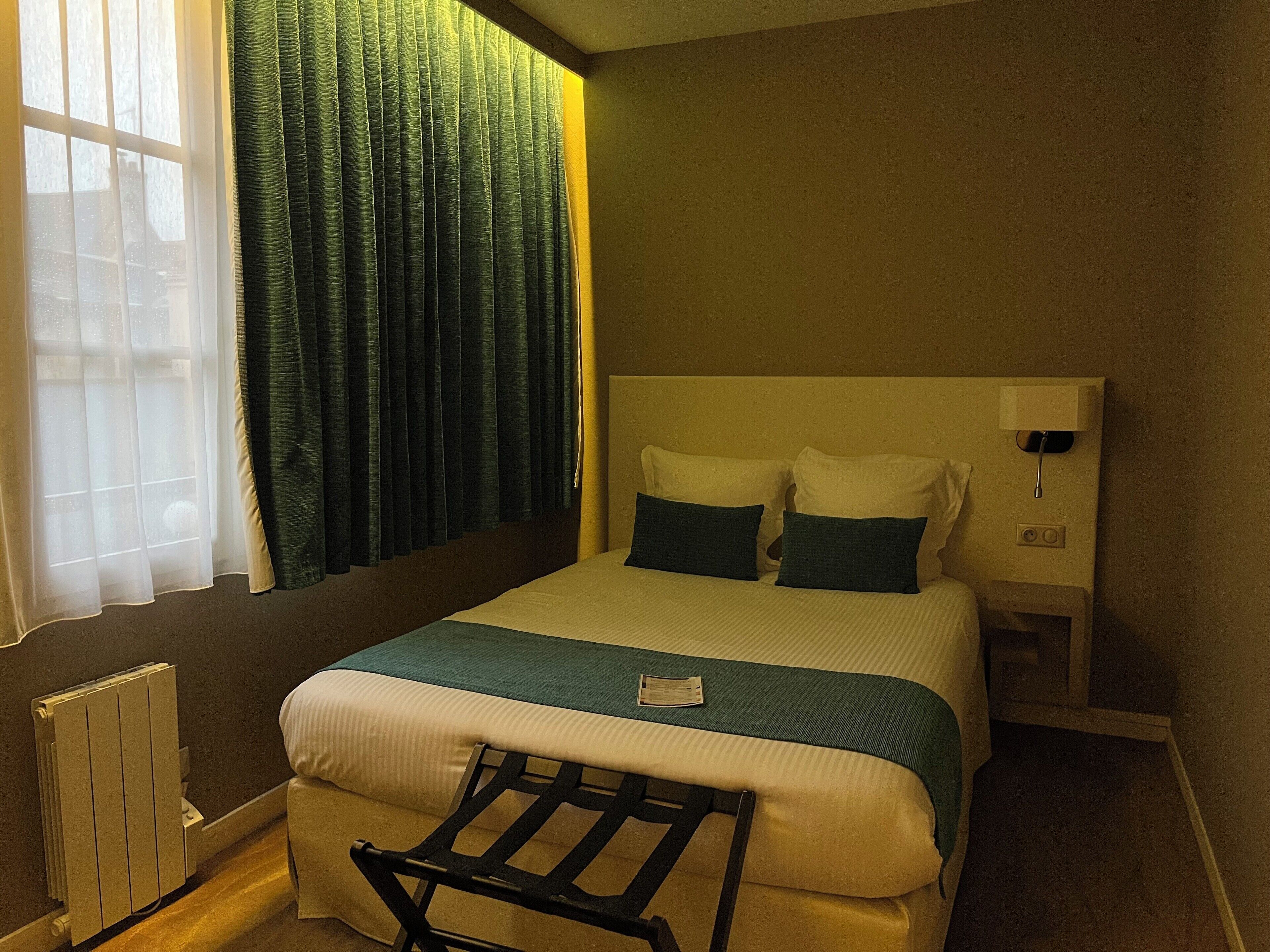 Ein Hotelzimmer mit Bett, Nachttisch, Lampe, Heizkörper und einem Fenster mit Vorhängen.