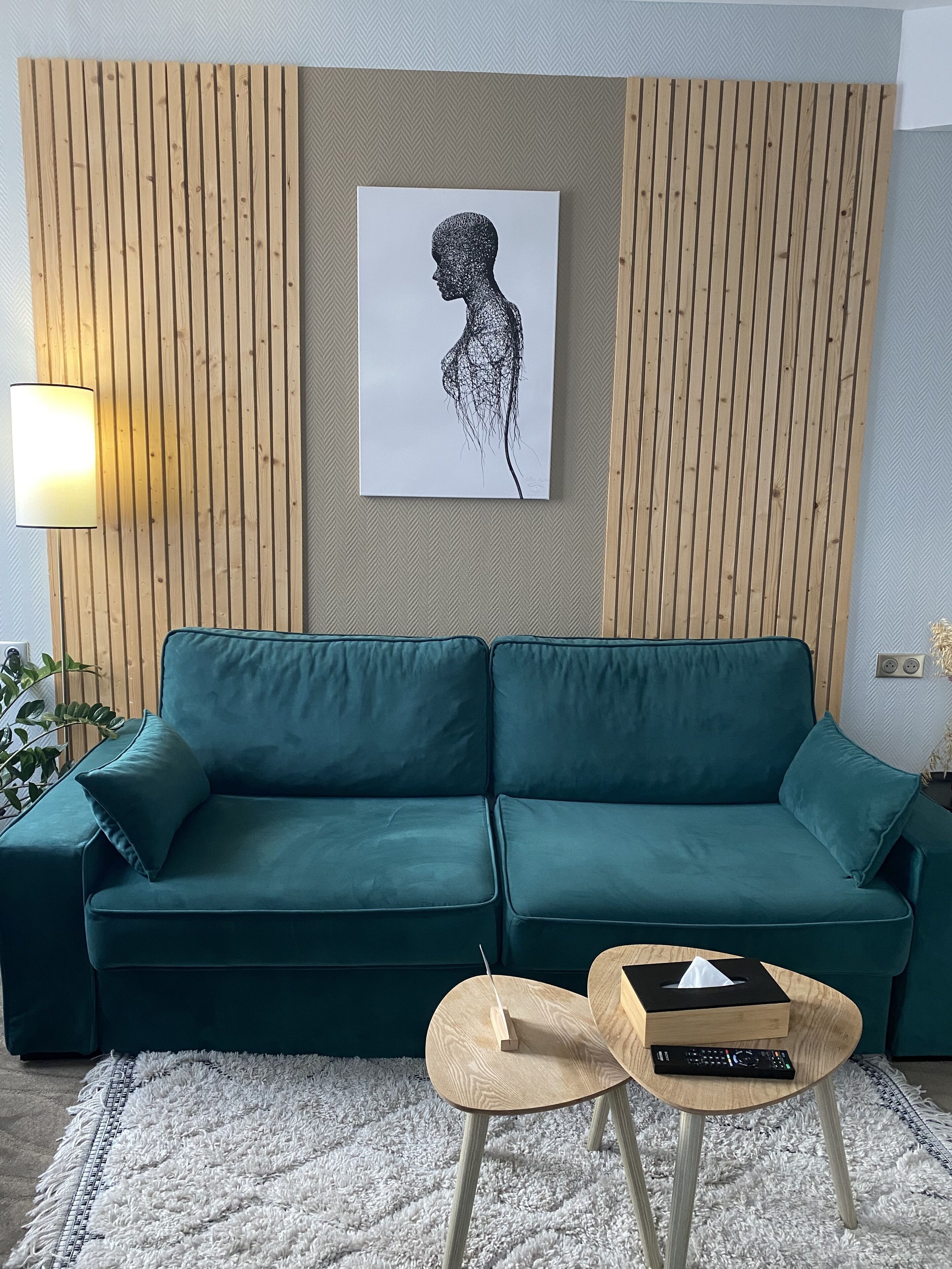 A living room with a teal sofa, a white rug, a wooden wall panel, and a framed artwork.