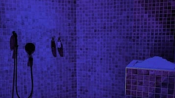 Área de duche com azulejos, parede revestida a azulejos, chuveiro e lavatório com dispensador de sabonete.