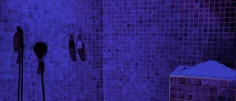 Un área de ducha con azulejos, pared azulejada, cabezal de ducha y lavamanos con dispensador de jabón.