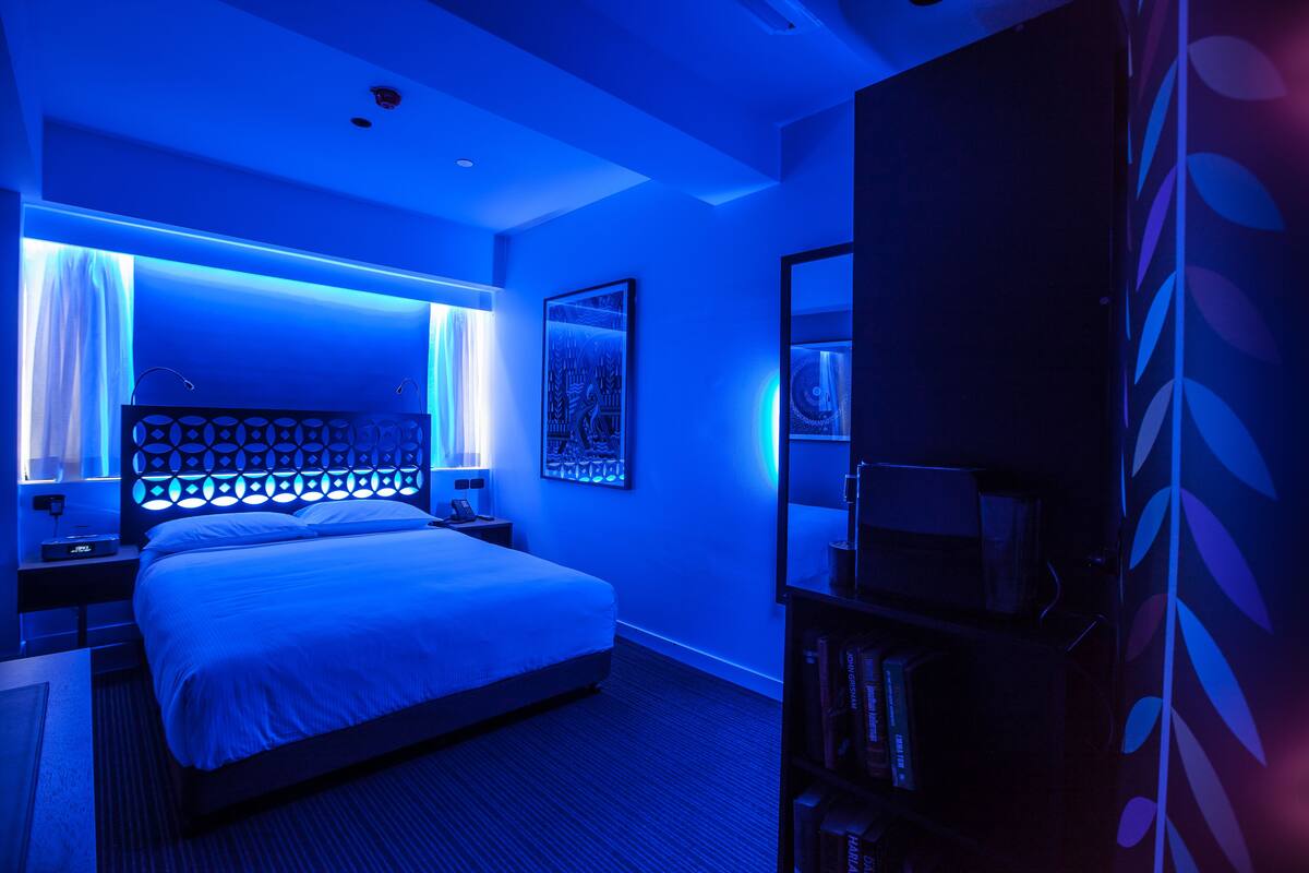 A modern hotel room with a large bed, a headboard with a geometric pattern, a bedside table with a lamp, and a bookshelf.