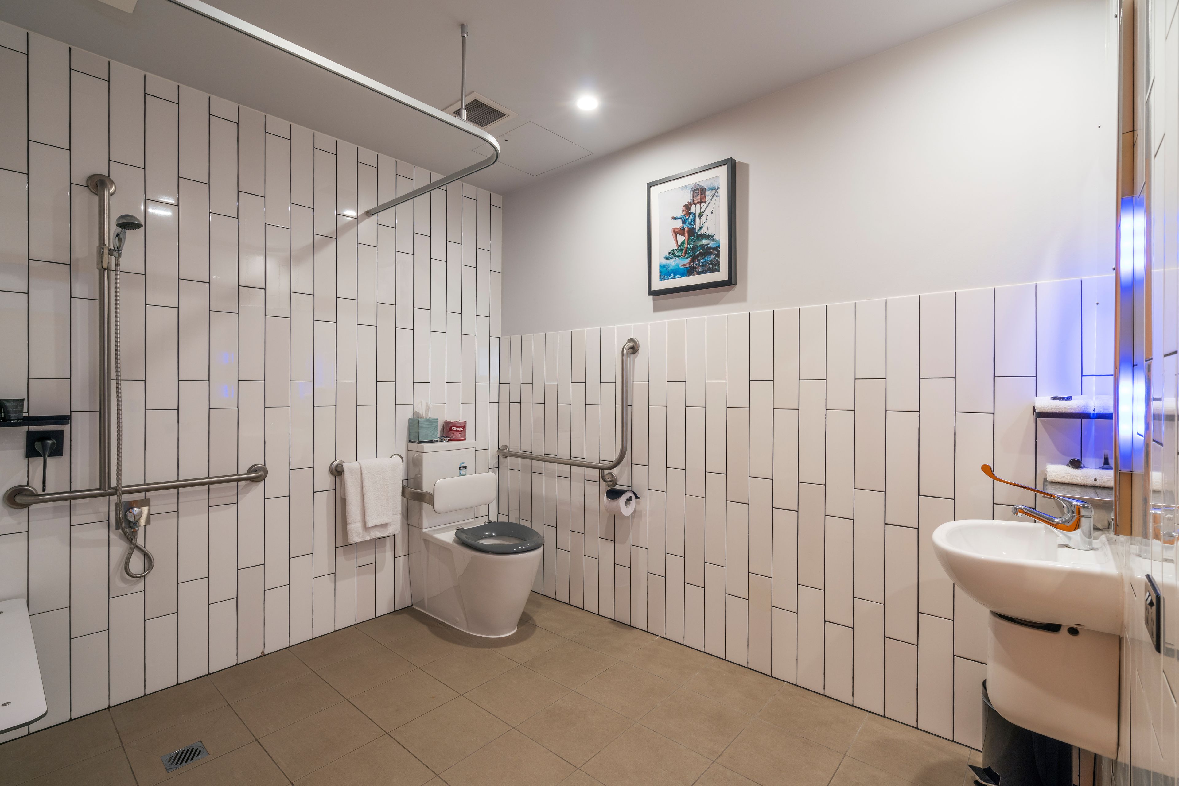 A modern bathroom with white tiled walls, a toilet with a grab bar, a shower area, and a sink with a mirror.