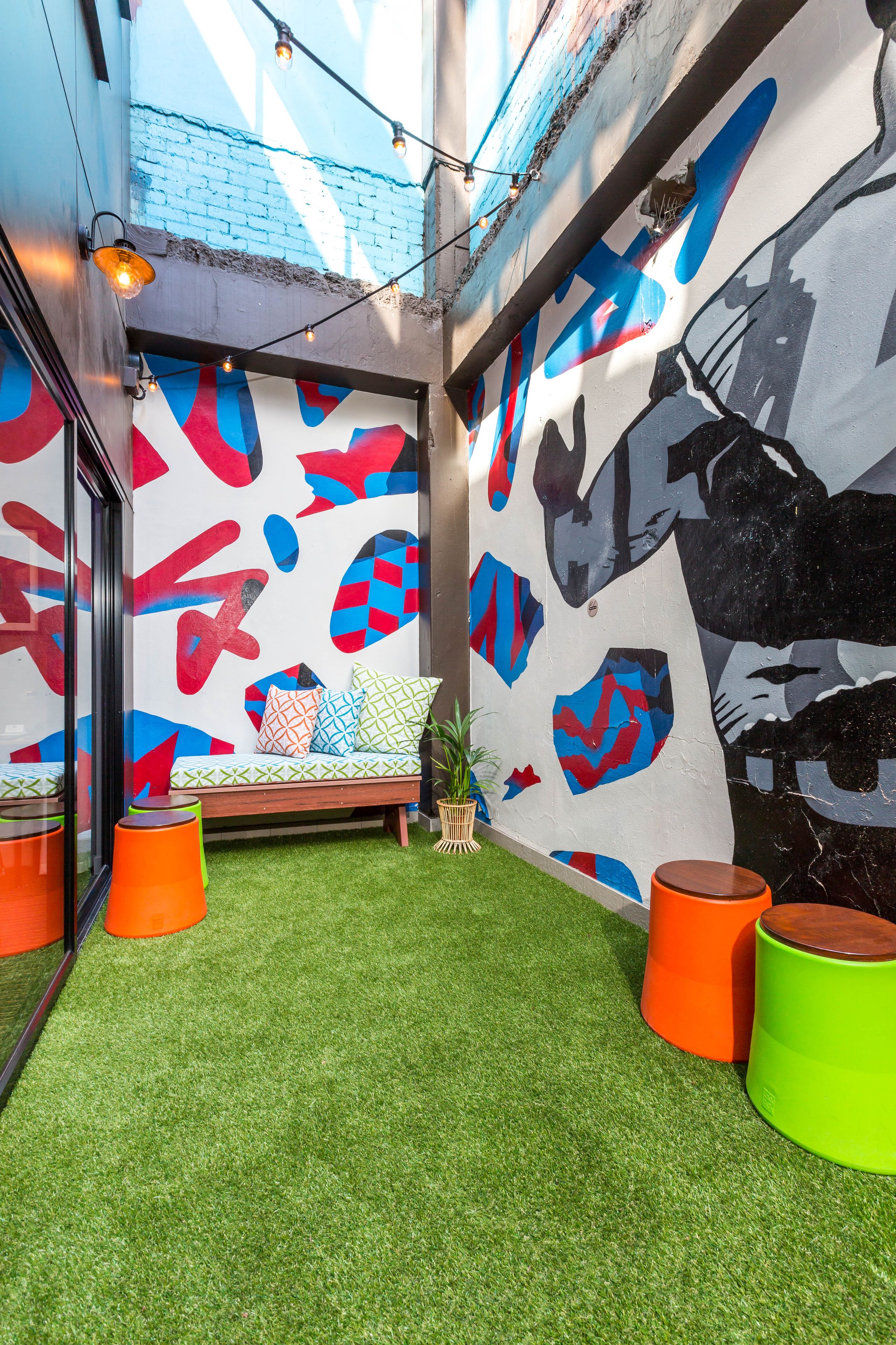 A modern outdoor seating area with artificial grass, colorful cushions, and a mural with abstract shapes.