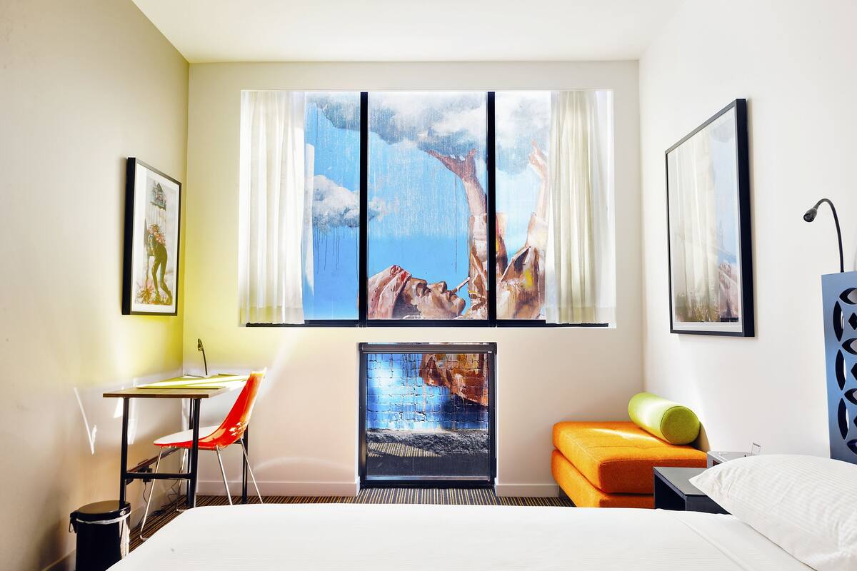 A hotel room with a bed, a desk with a chair, a window with a painting, and a sofa.