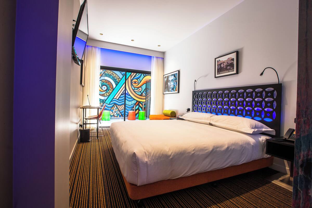 A modern hotel room with a large bed, a headboard with circular lights, a flat-screen TV, and a colorful abstract wall art.