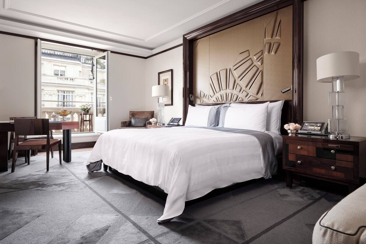 A hotel room with a large bed, a desk, a chair, a nightstand, and a view of the city through the window.