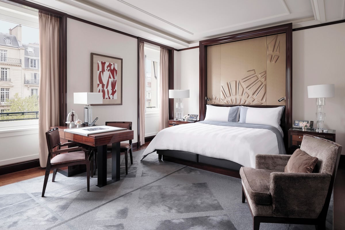A hotel room with a large bed, a desk with a chair, a dining table with chairs, and a view of the city through the window.