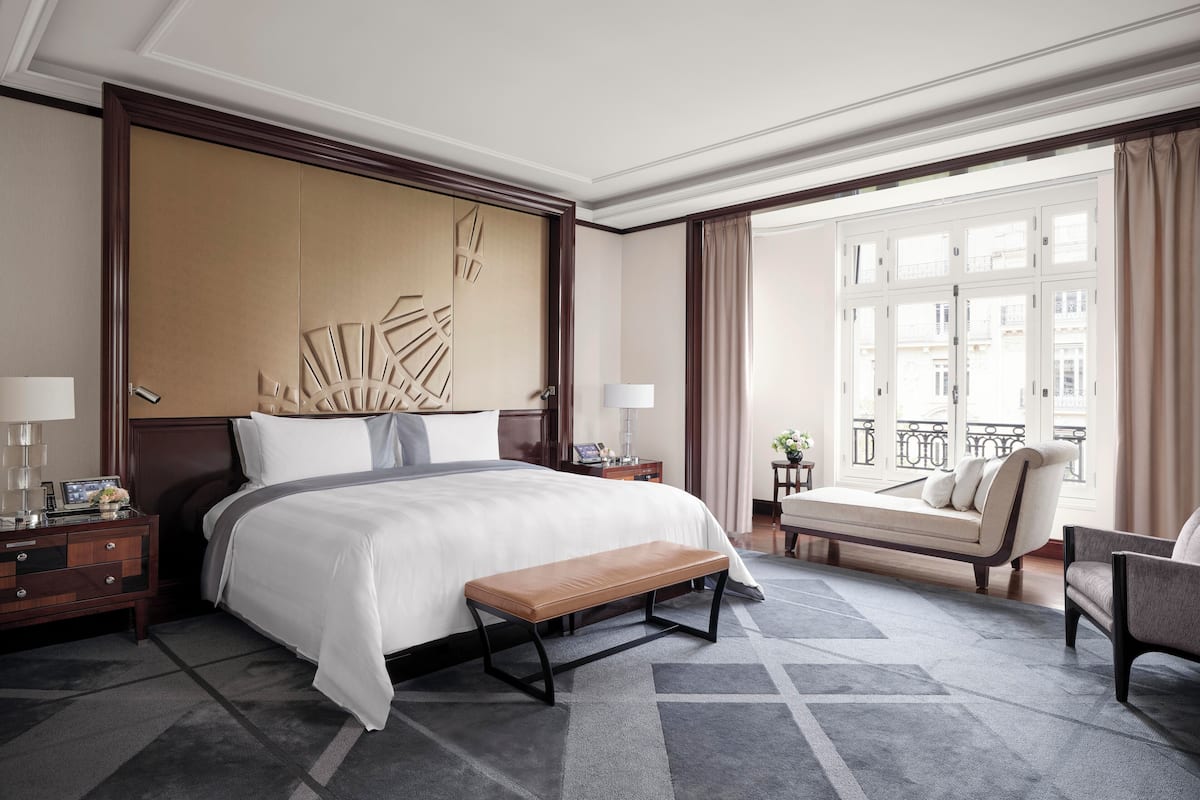 A hotel room with a large bed, a bench, a sofa, and a view of the city through large windows.