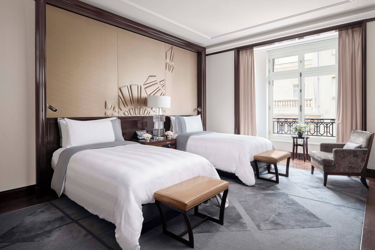 A hotel room with two beds, a wooden bench, a desk with a lamp, and a view of a balcony.