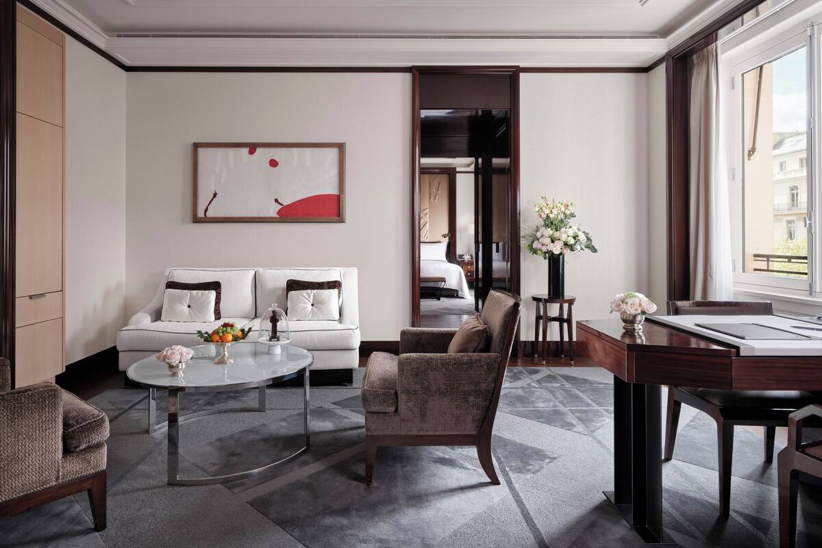 A modern hotel room with a sofa, armchair, dining table, and a view of the city through large windows.