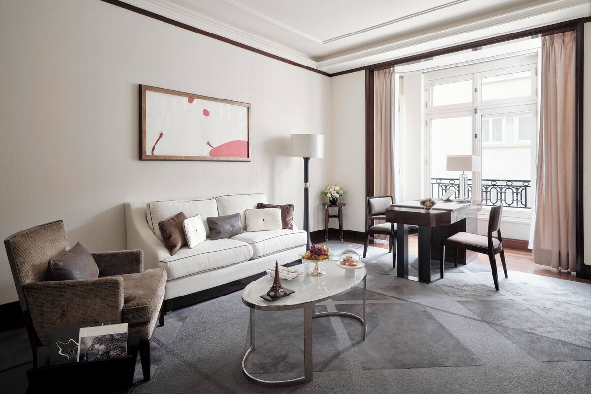 A modern hotel room with a sofa, armchair, coffee table, and a dining area with a table and chairs.