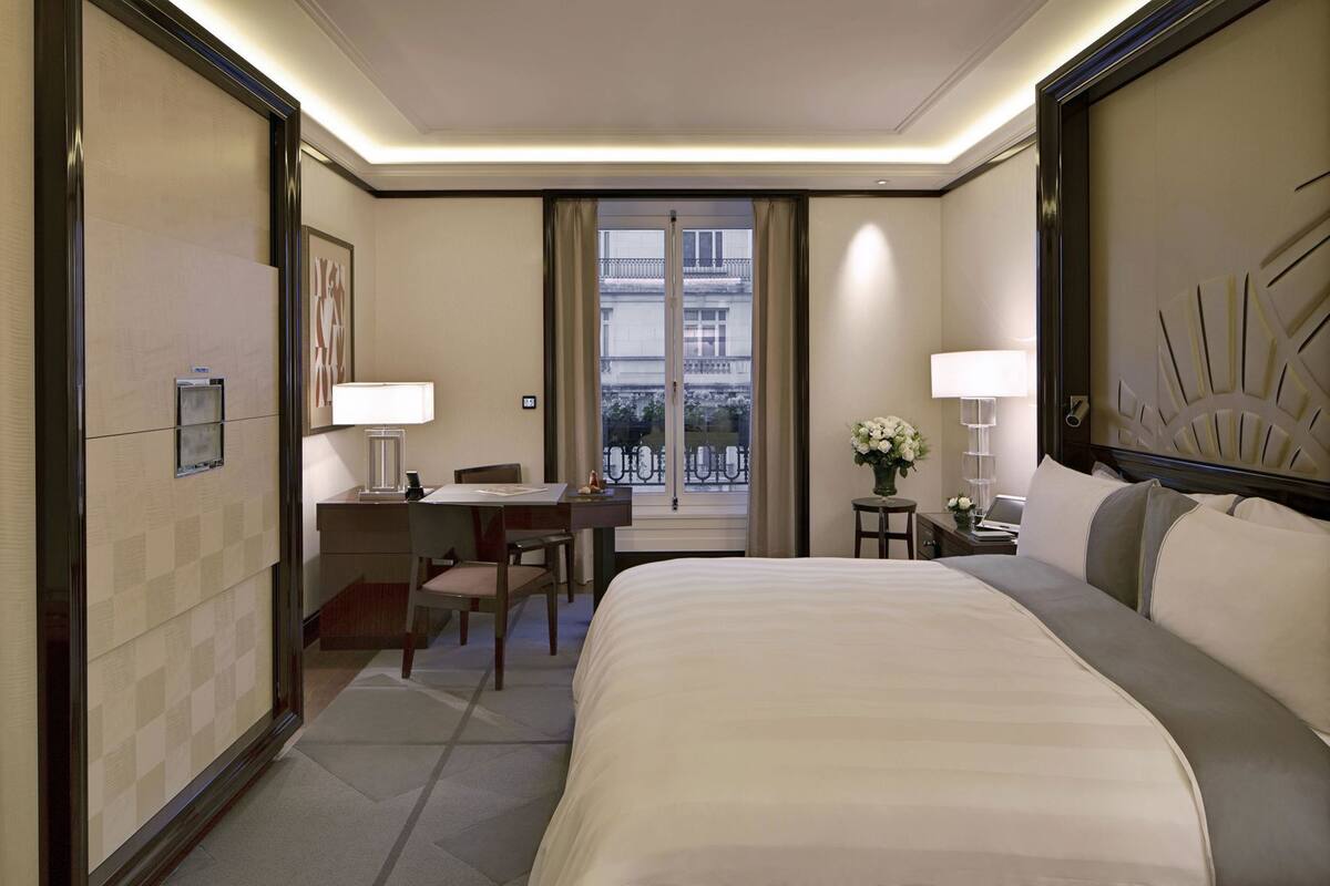 A hotel room with a bed, desk, chair, and a view of the city through the window.