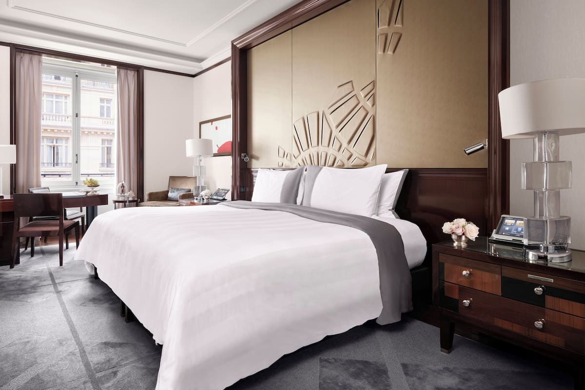 A hotel room with a large bed, a desk with a chair, a bedside table with a lamp, and a view of buildings through the window.