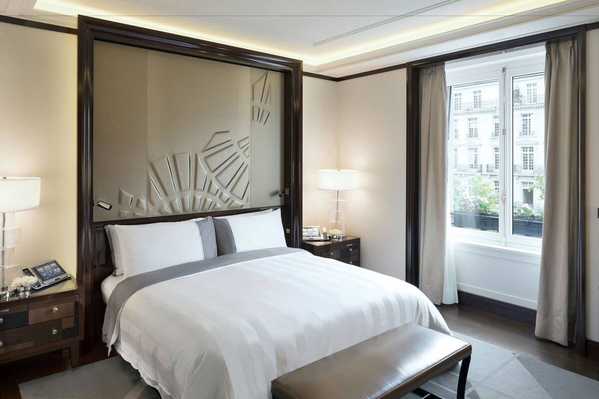 A hotel room with a large bed, bedside tables, a bench, a decorative headboard, and a window with a view of buildings.