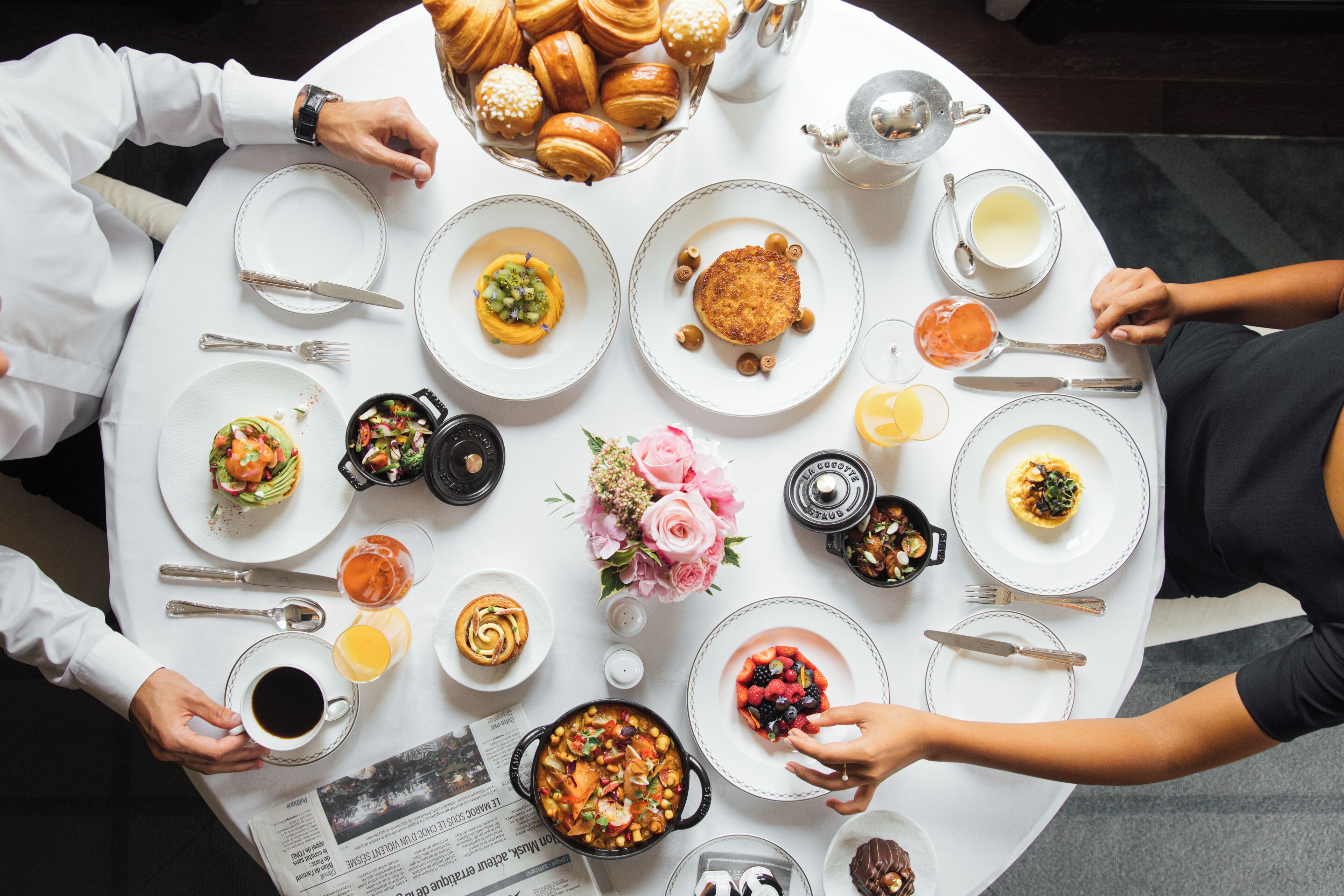Una mesa redonda de comedor con diferentes platos, un ramo de flores y un periódico.