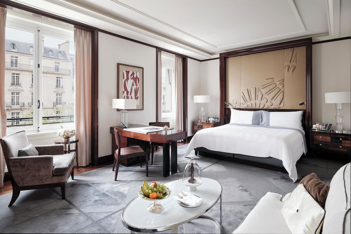 A hotel room with a large bed, a desk with a chair, a dining table with fruit, and a view of buildings through the window.