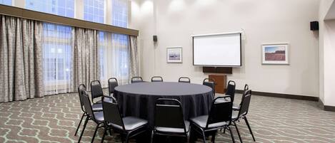 Un salón de conferencias con una mesa redonda, sillas negras, una pantalla de proyector y dos cuadros en la pared.