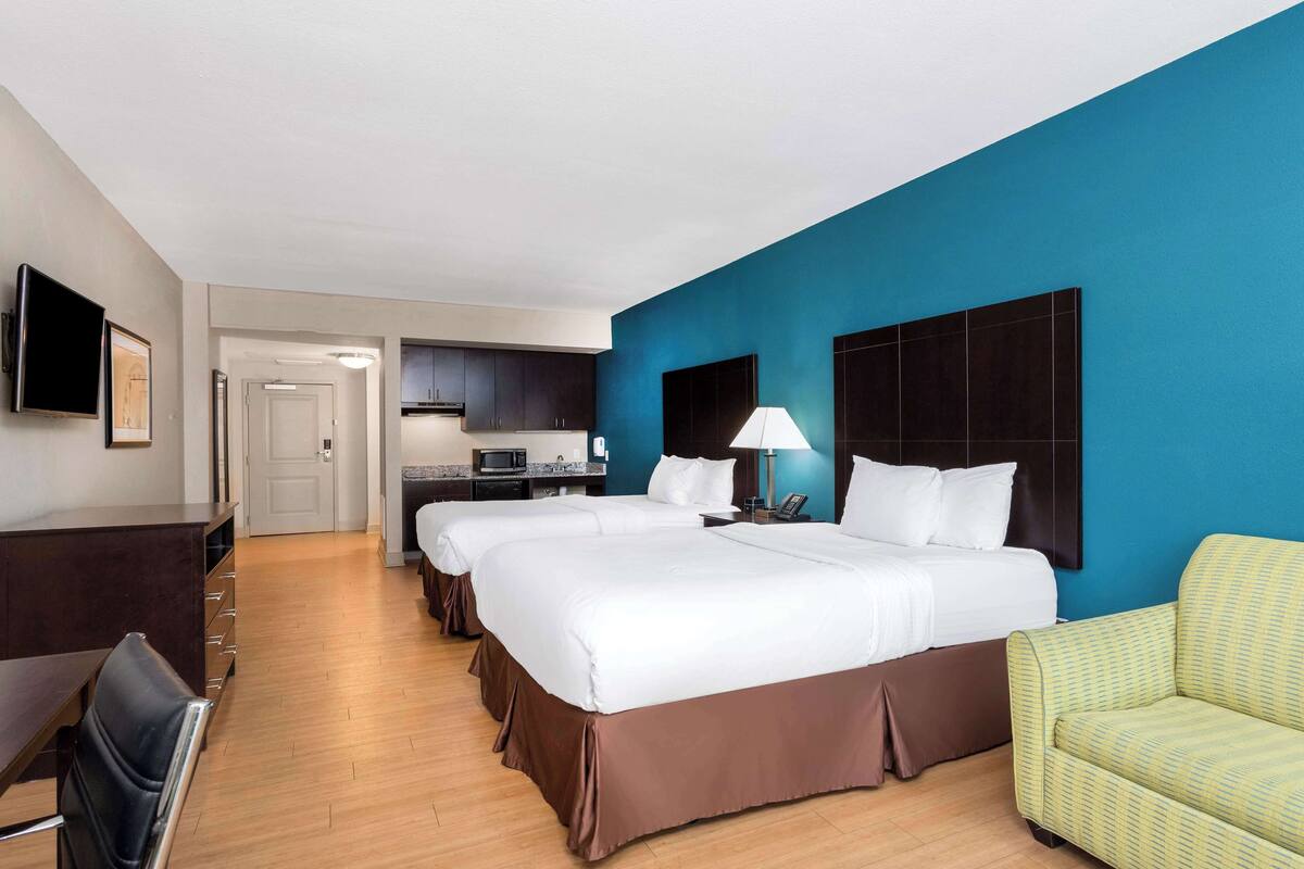 Une chambre d’hôtel avec deux lits, un bureau, une chaise, une télévision, un coin cuisine et un mur d’accent bleu.