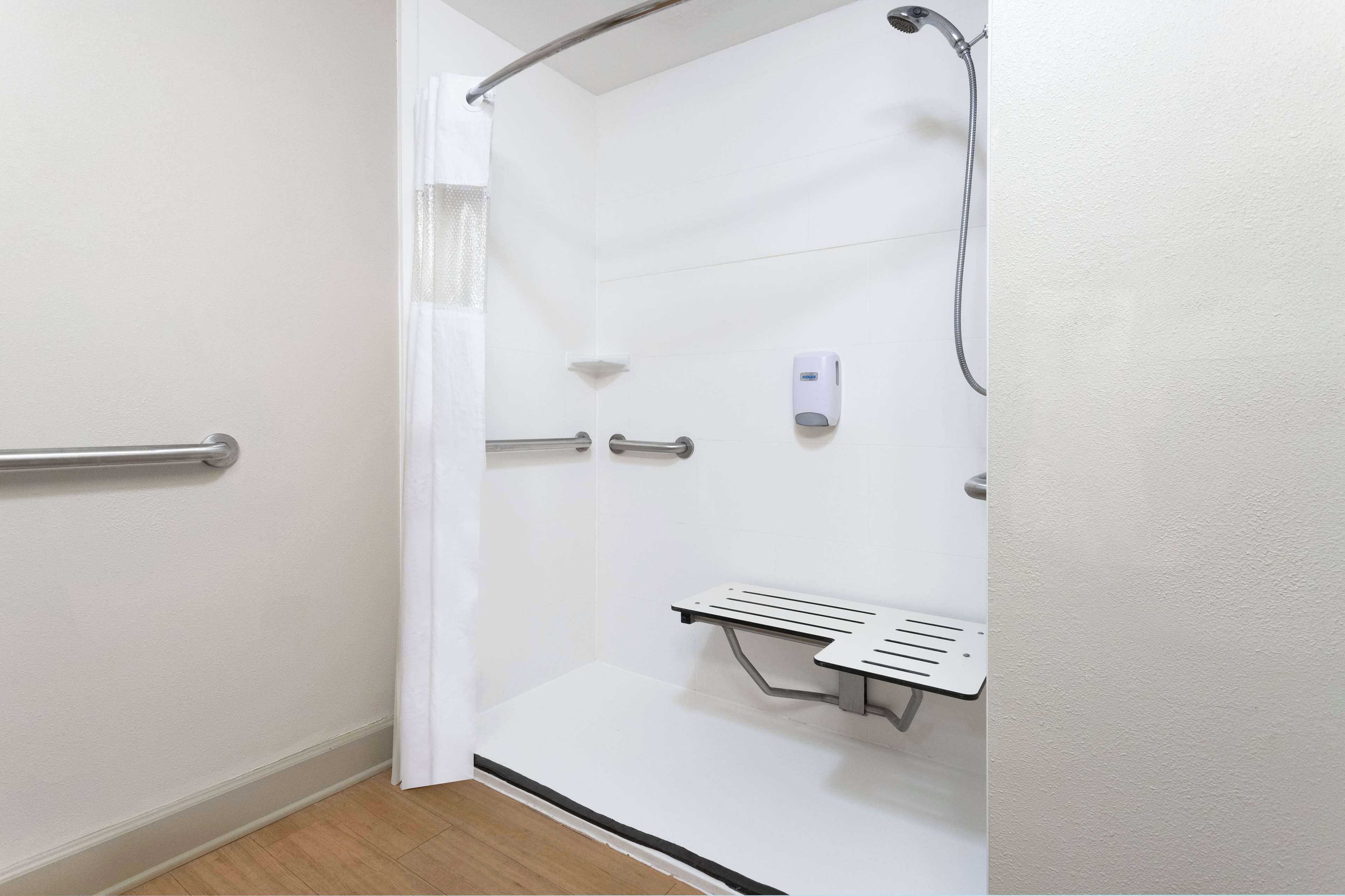 A modern bathroom with a shower, handrails, a soap dispenser, and a bench.