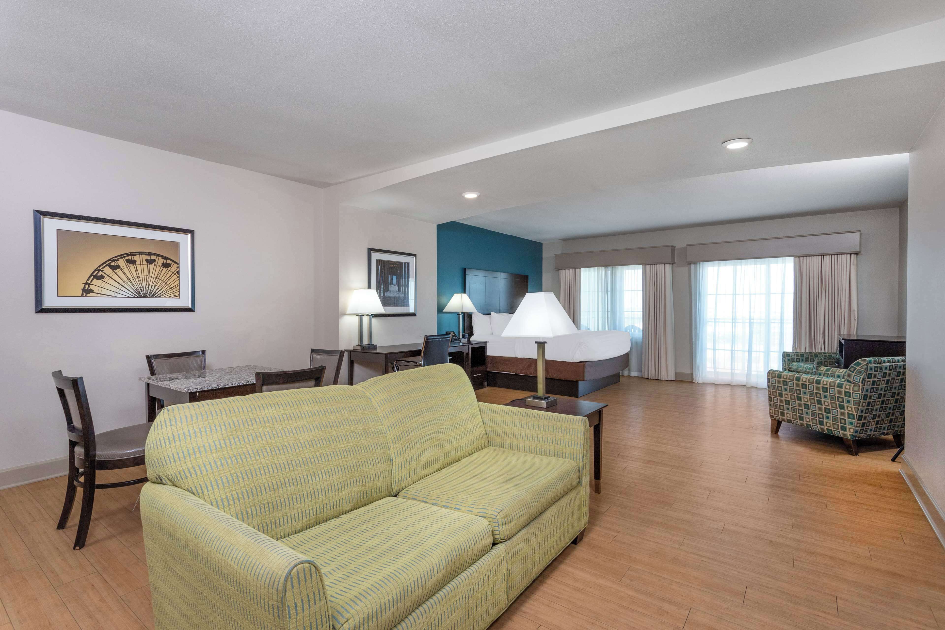 A hotel room with a green sofa, a dining table with chairs, a TV, and a bed with curtains.