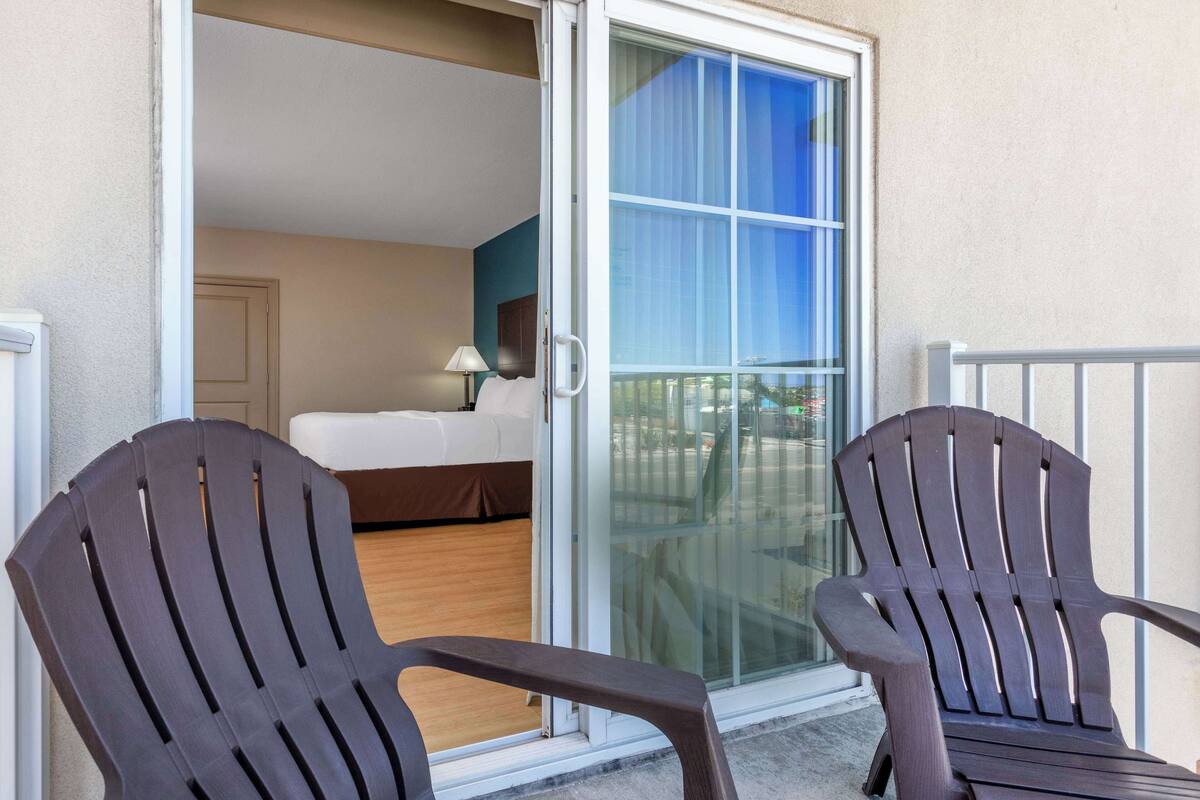 Un balcon avec deux fauteuils Adirondack, une porte-fenêtre coulissante donnant sur une chambre à coucher équipée d’un lit et d’une table de chevet, et une vue sur l’extérieur.