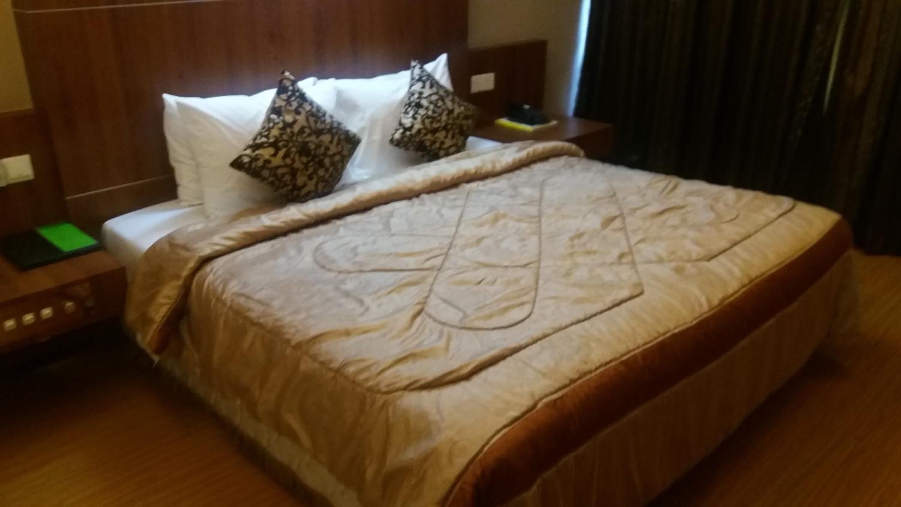 A hotel room with a large bed, a wooden headboard, two patterned pillows, a green book on the nightstand, and a wooden floor.