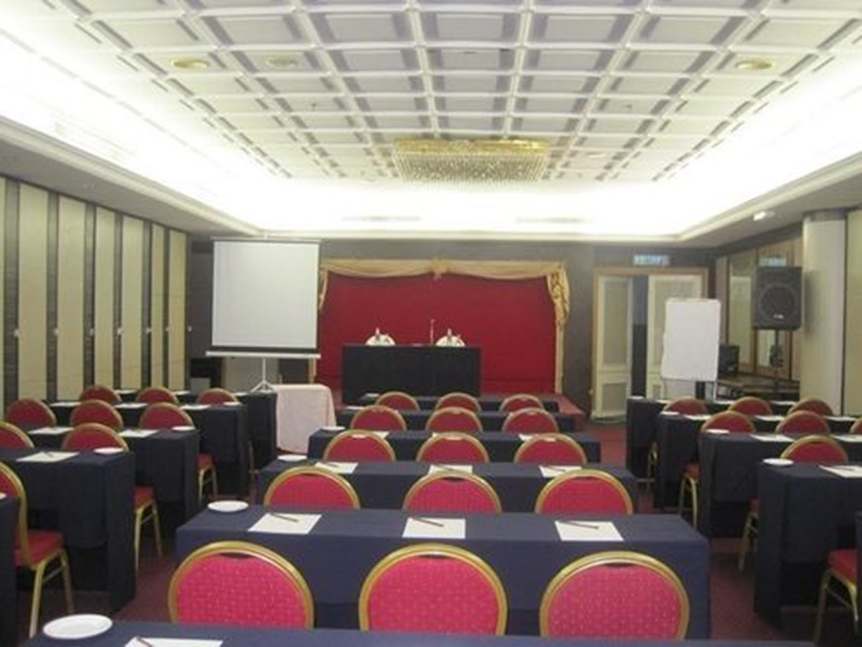 A conference room with rows of tables and chairs, a podium, a whiteboard, and a stage with a red curtain.