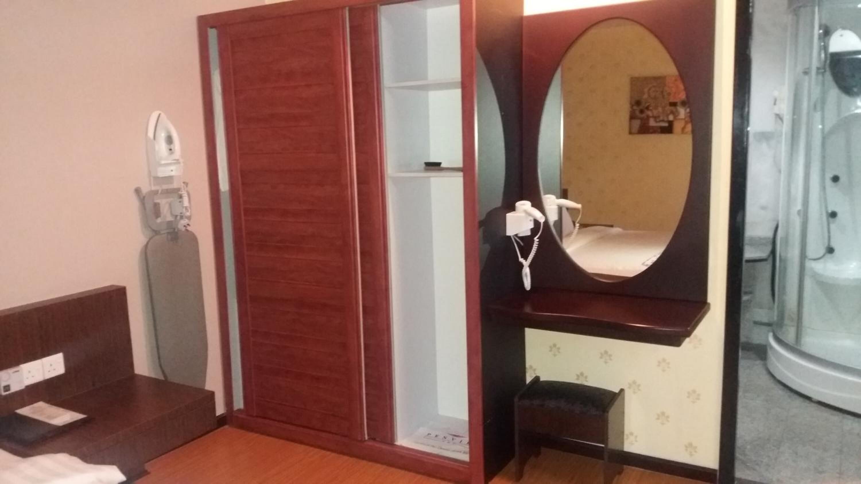 A hotel room with a wooden wardrobe, a vanity with a mirror, a hairdryer, and a shower area.