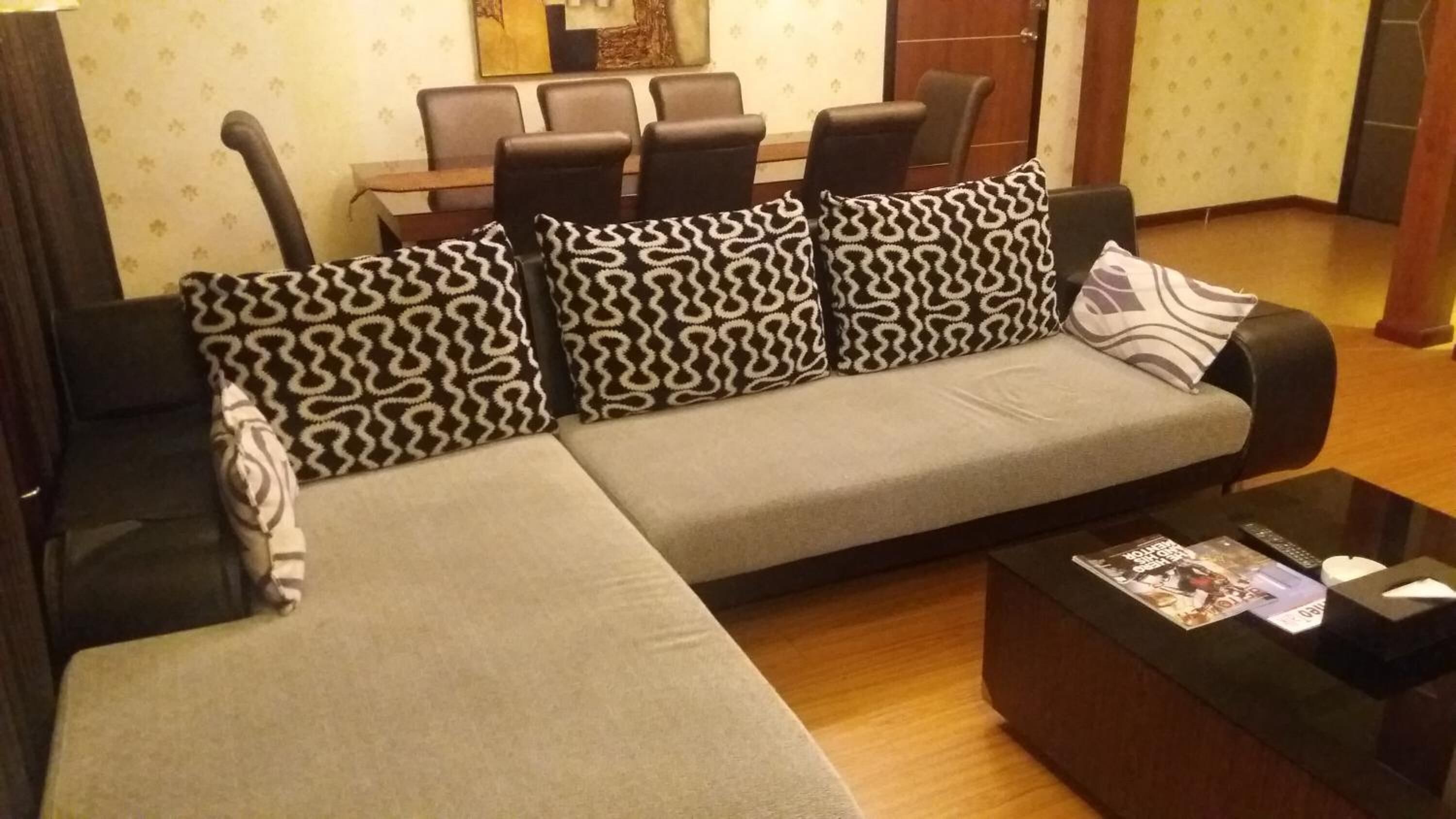 Un salon moderne avec un canapé gris, des coussins à motifs, une table basse avec des magazines et, au fond, un coin repas.