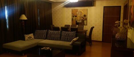 Uma sala de estar com um sofá seccional, uma mesa de centro, uma luminária, uma área de jantar com cadeiras e um quadro na parede.