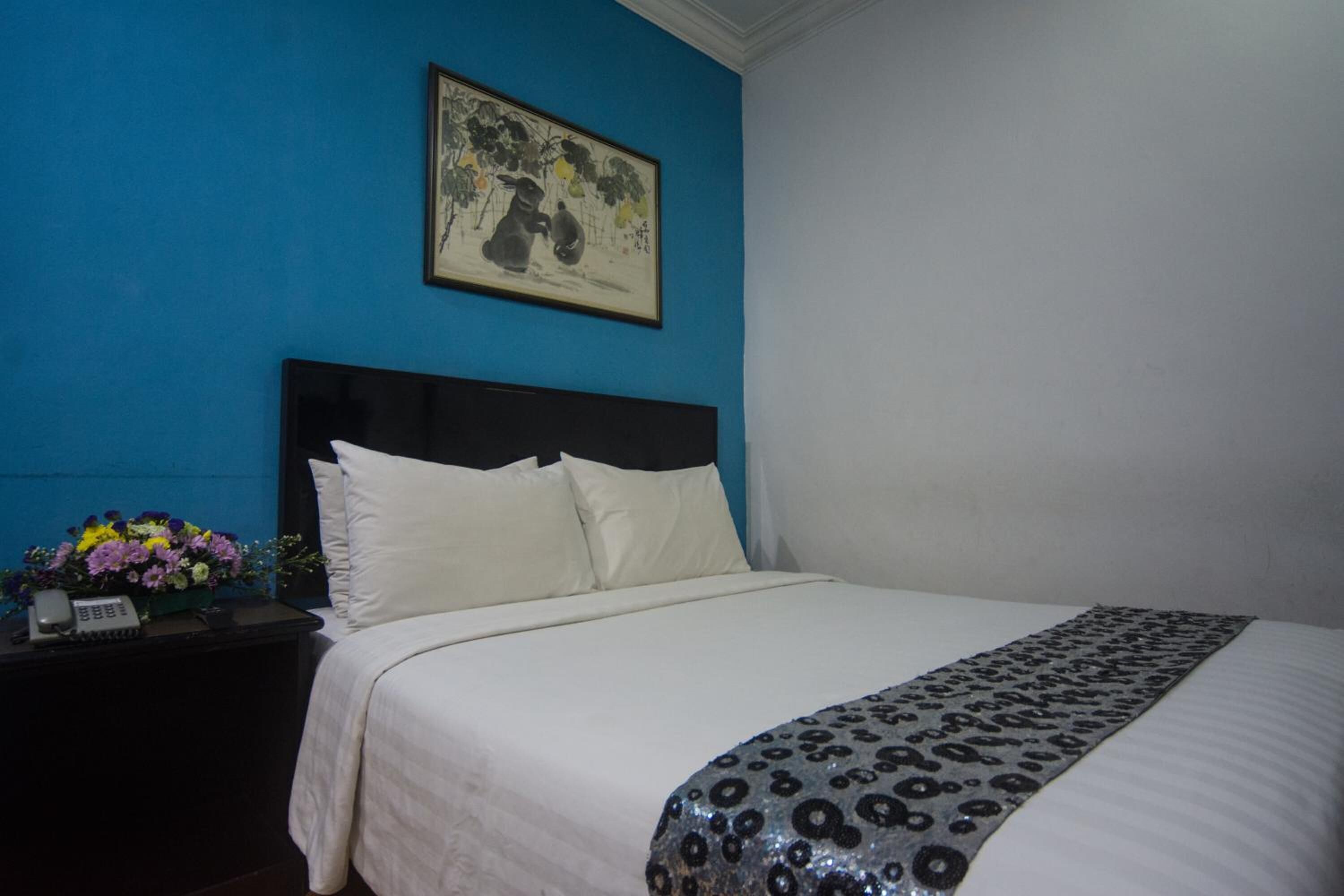 A hotel room with a bed, white bedding, a blue wall, a framed artwork, and a nightstand with a phone and flowers.