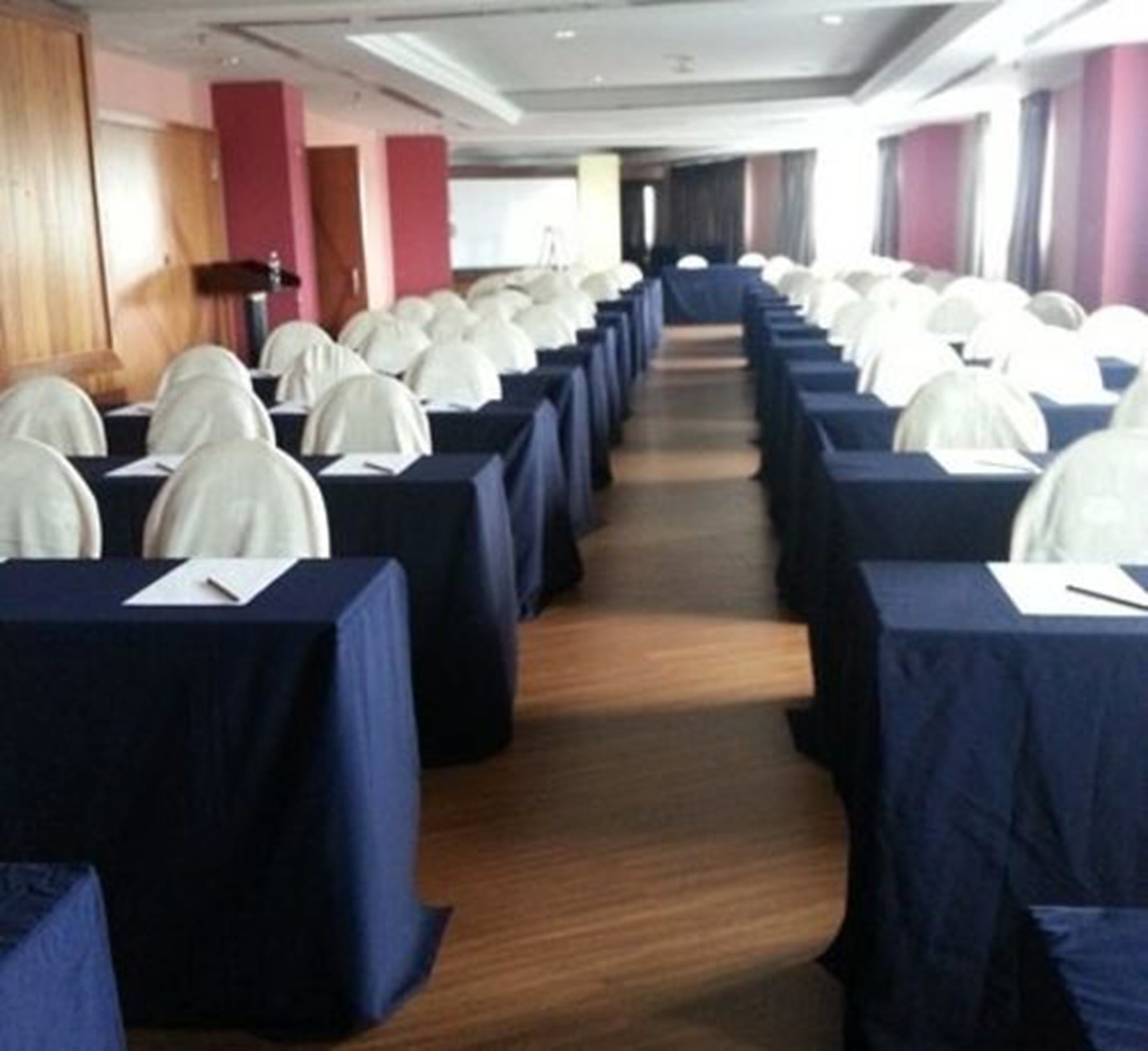 A conference room with rows of tables and chairs, a wooden floor, and a view of the hallway.
