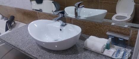 Un baño con lavamanos blanco, espejo, inodoro y toallero con artículos de aseo.