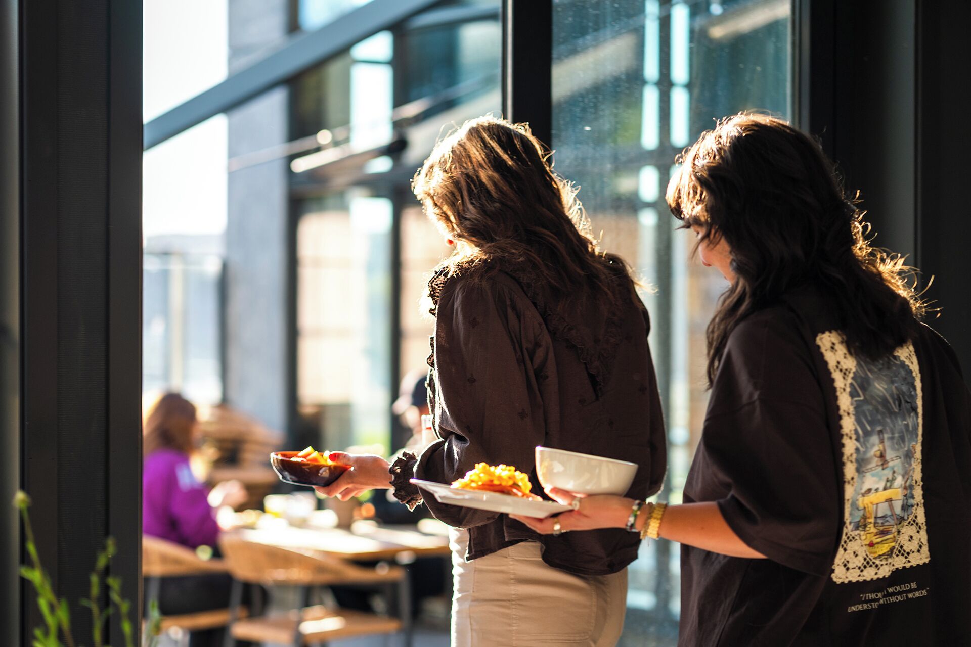 Dos personas en un ambiente interior moderno con ventanas amplias, sosteniendo platos de comida.