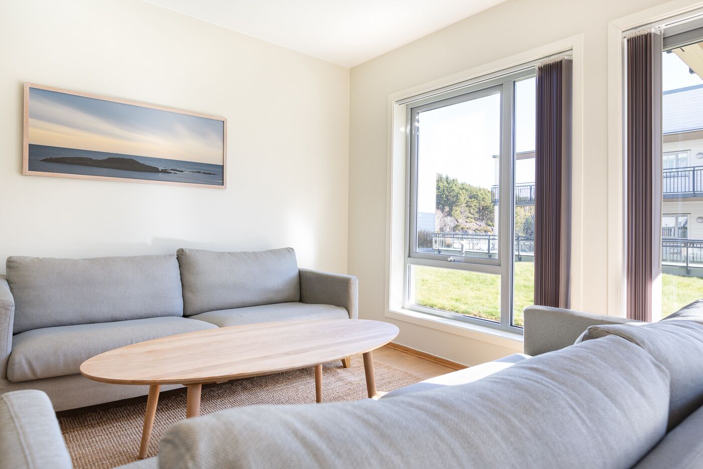 A modern living room with a grey sofa, a wooden coffee table, and a large window offering a view of a building and greenery.
