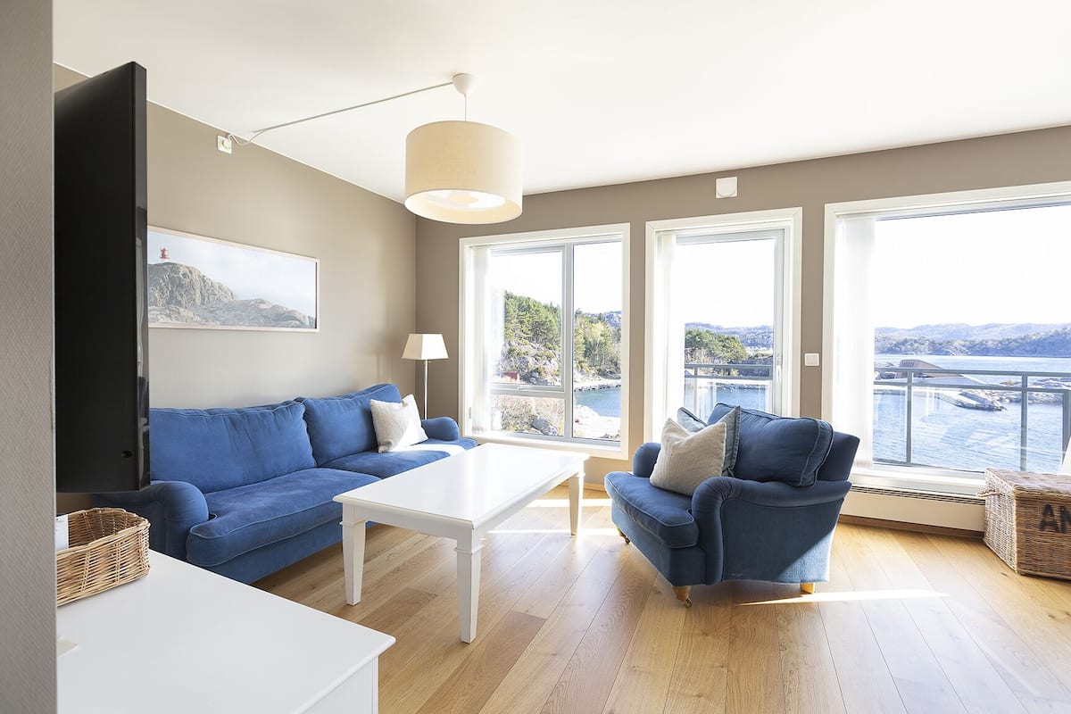 A modern living room with a blue sofa, a white coffee table, and a blue armchair. There is a large window offering a view of a body of water and hills.