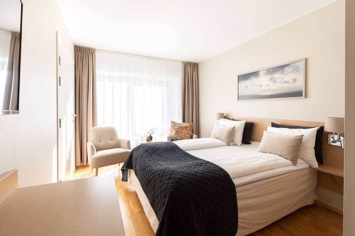 A hotel room with a large bed, a chair, a desk, and a window with curtains.