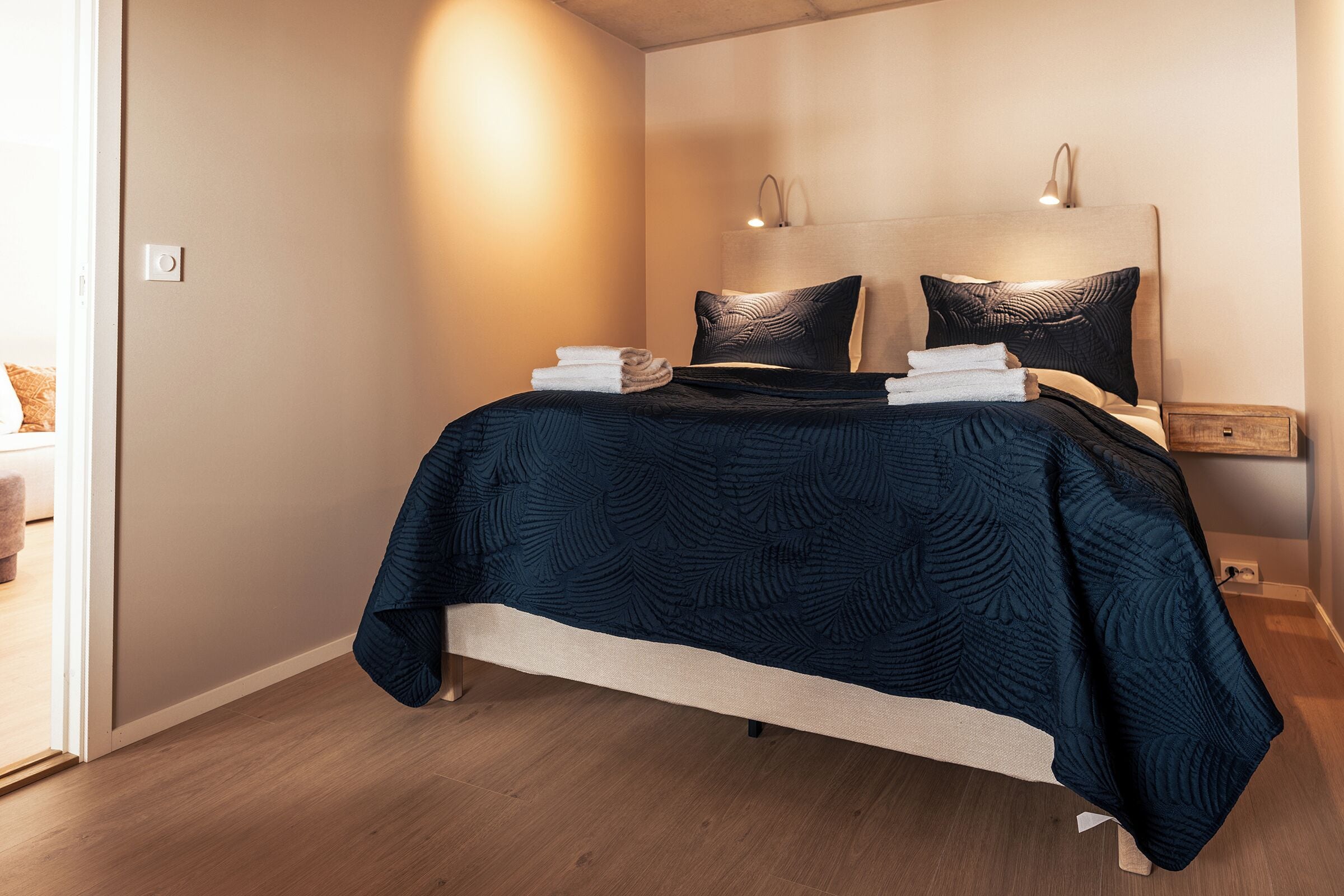 Una cama bien hecha con un edredón oscuro estampado, dos almohadas y toallas dobladas sobre un suelo de madera.