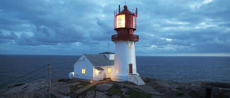 Ein Leuchtturm mit weißem Unterteil und roter Spitze, ein kleines Nebengebäude und eine felsige Küstenlinie mit dem Meer im Hintergrund.