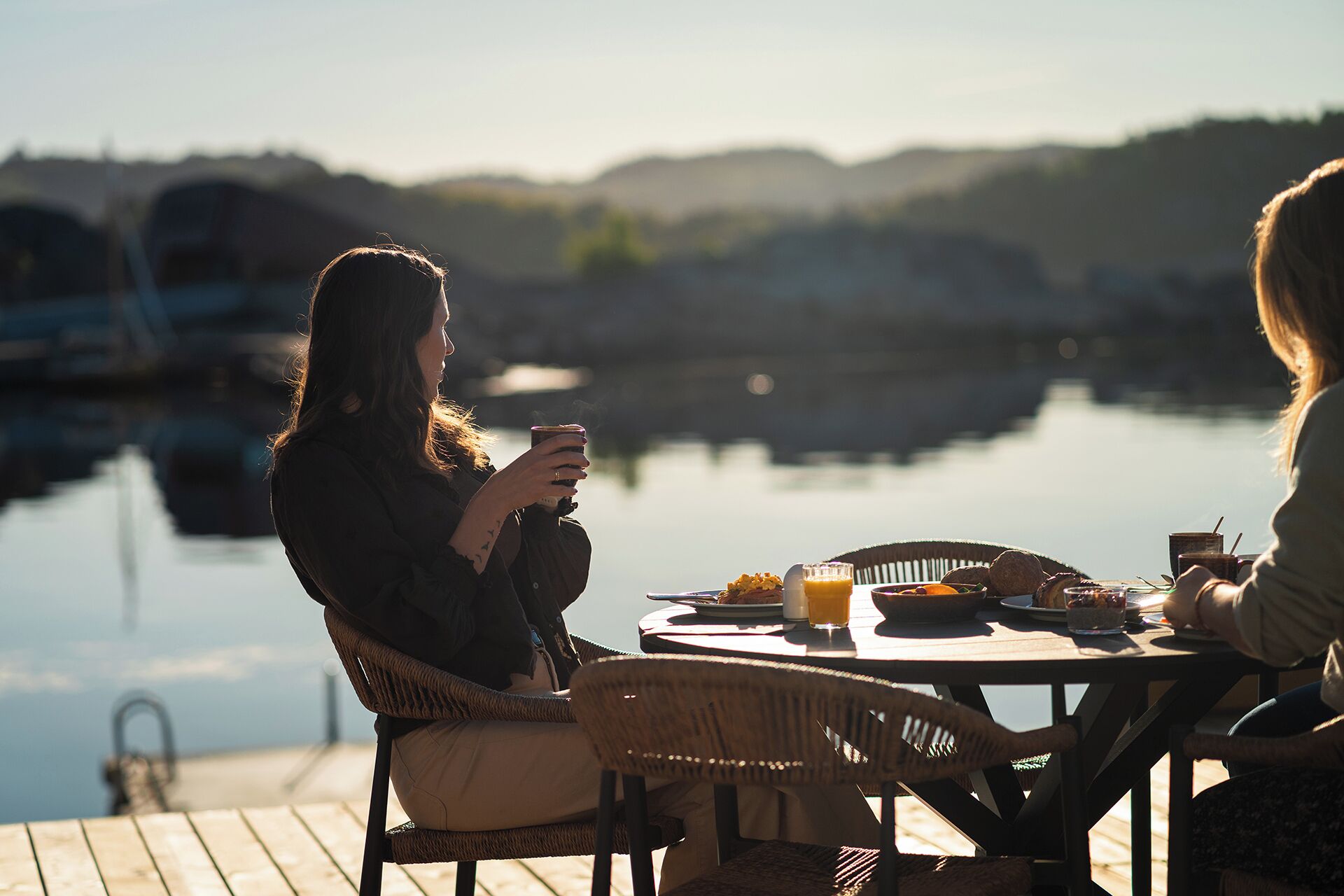 A dining table with chairs, a person holding a cup, and a view of a body of water with boats in the background.