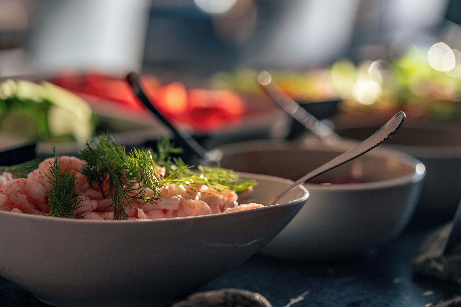 Eine Schüssel mit Garnelen, dekoriert mit Dill, einem Löffel und einem unscharfen Hintergrund mit weiteren Speisen.