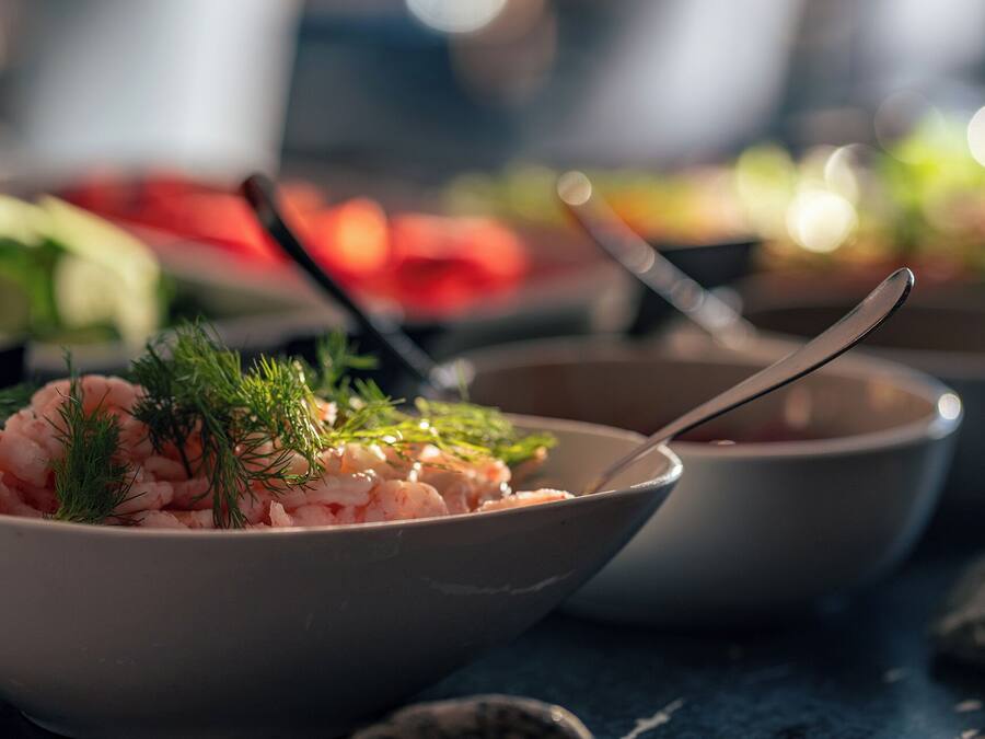 A bowl of shrimp garnished with dill, a spoon, and a blurred background of other food items.