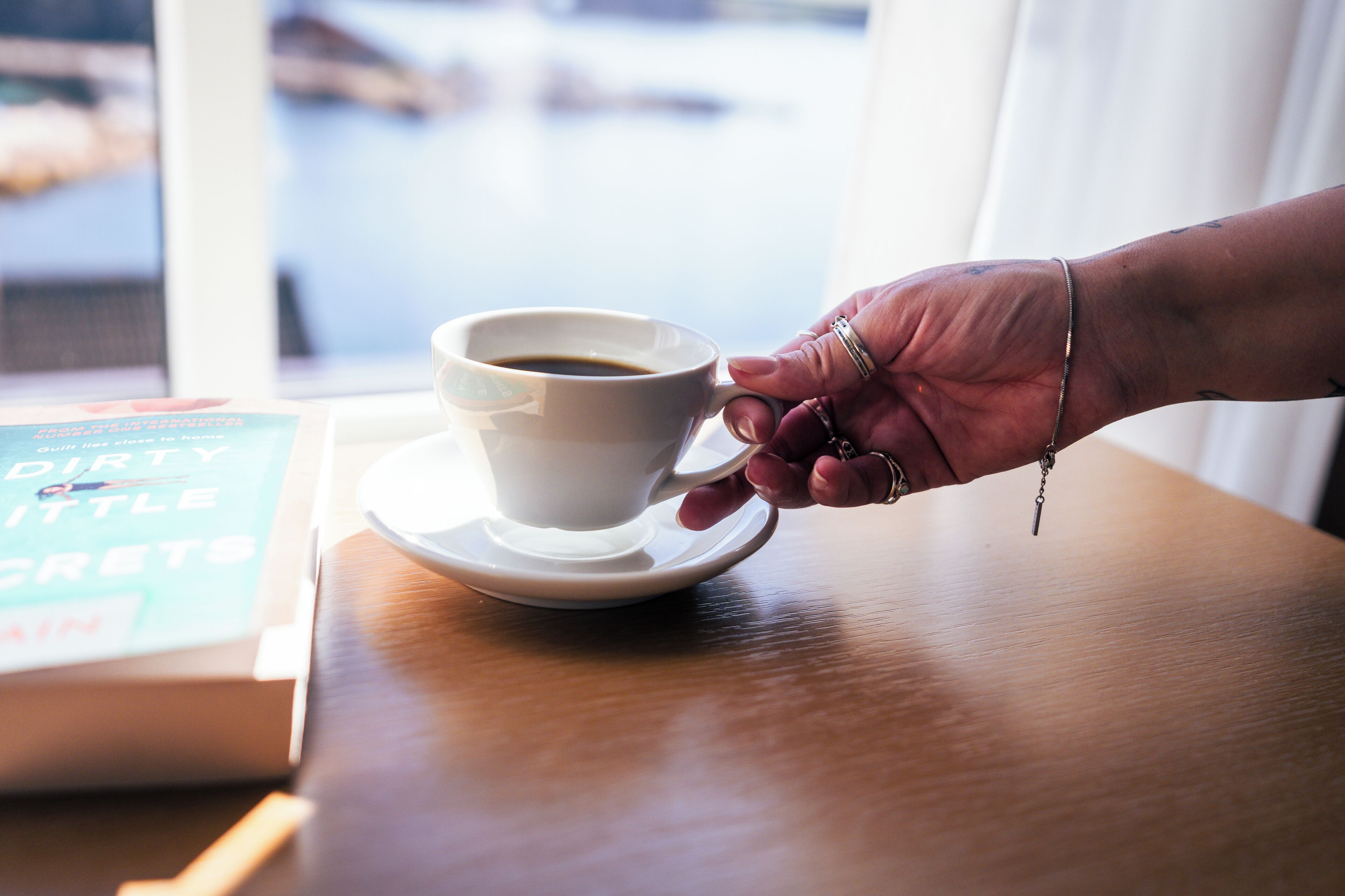 Eine Hand hält eine weiße Kaffeetasse auf einem Untertasse, auf einem Tisch liegt ein Buch mit dem Titel 'Dirty Little Secrets' und ein Fenster bietet Blick auf ein Gewässer.