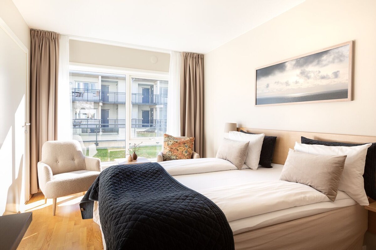 Una habitación de hotel con una cama, una silla, un escritorio, una ventana con vistas al exterior y un cuadro en la pared.